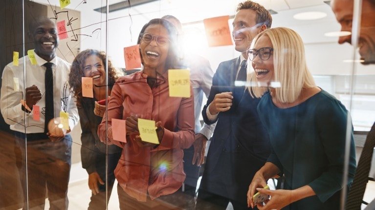 6 co-workers laughing in front of a clear wall that has post-it notes on it