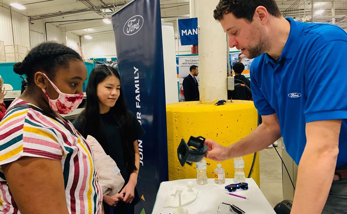 Ford employee showing 2 students an automative part at a local Ford event